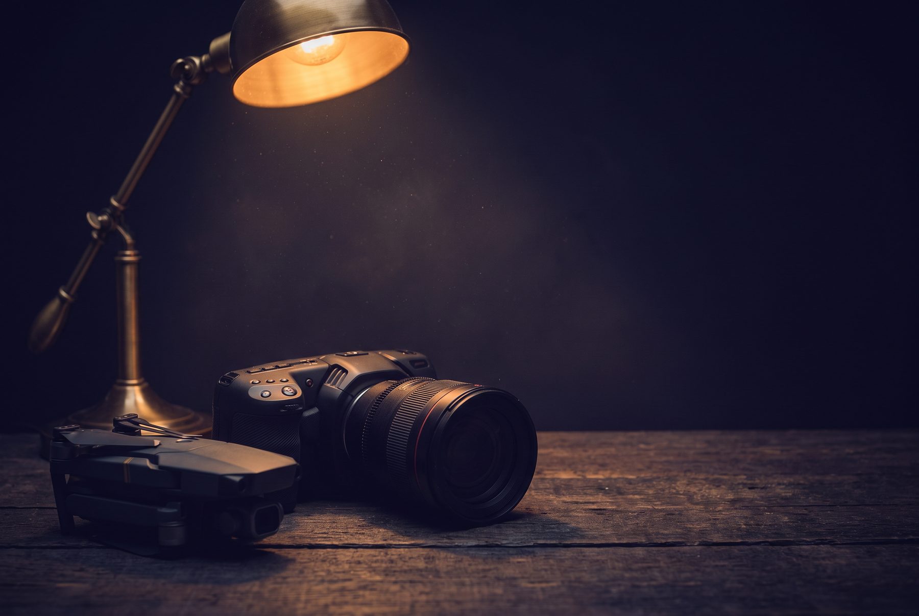 A cinema camera and a folded drone on a dark table under a warm lamp