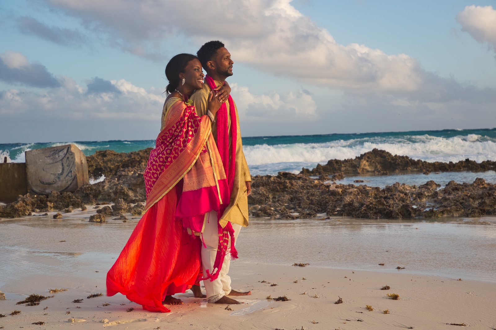 Couple in a red sari and gold kurta walking a rocky beach at sunset