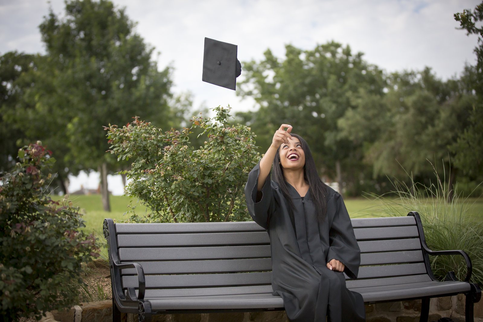 Graduate on a park bench tossing a mortarboard into the air