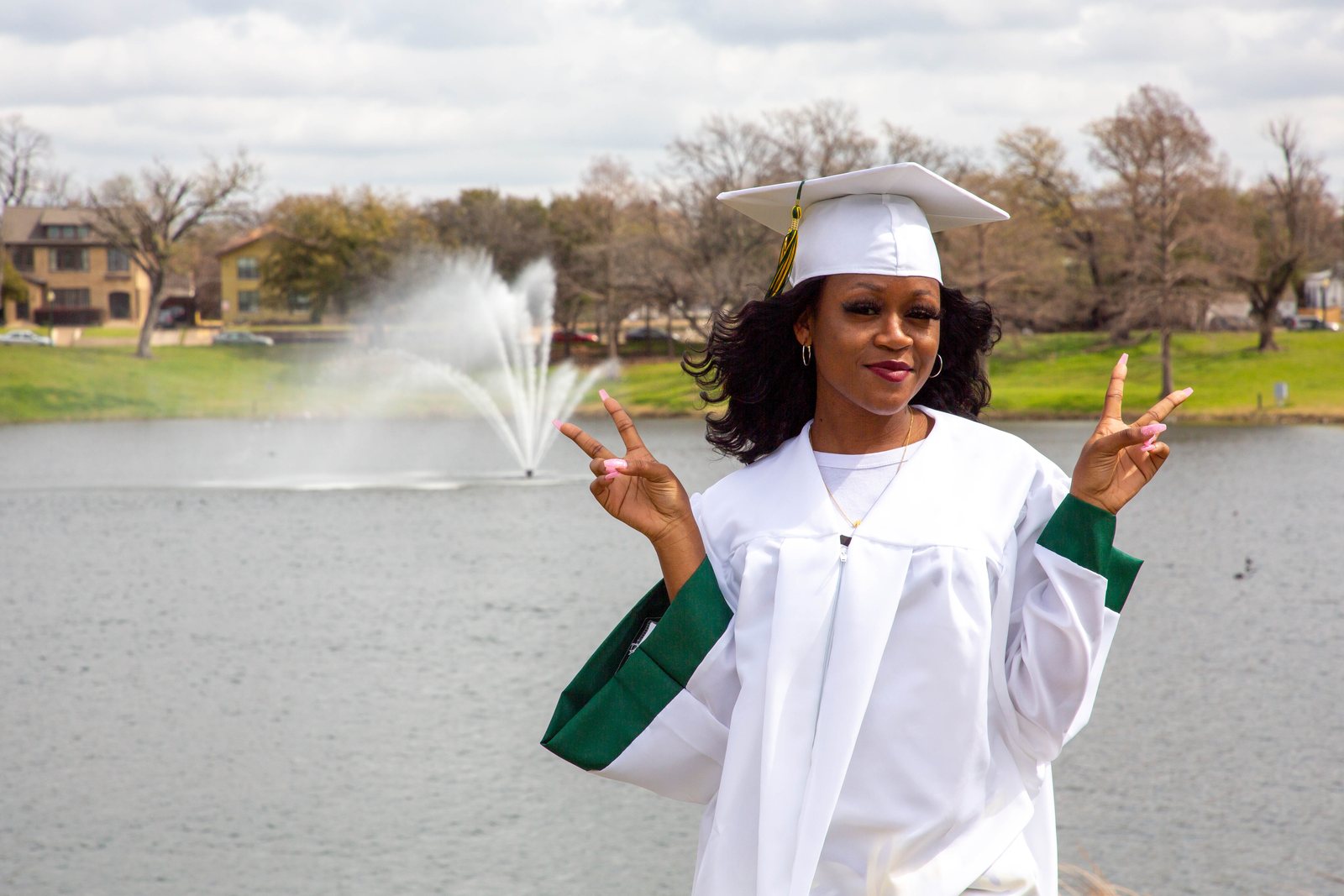 Graduate in a white gown flashing peace signs in front of a lake fountain