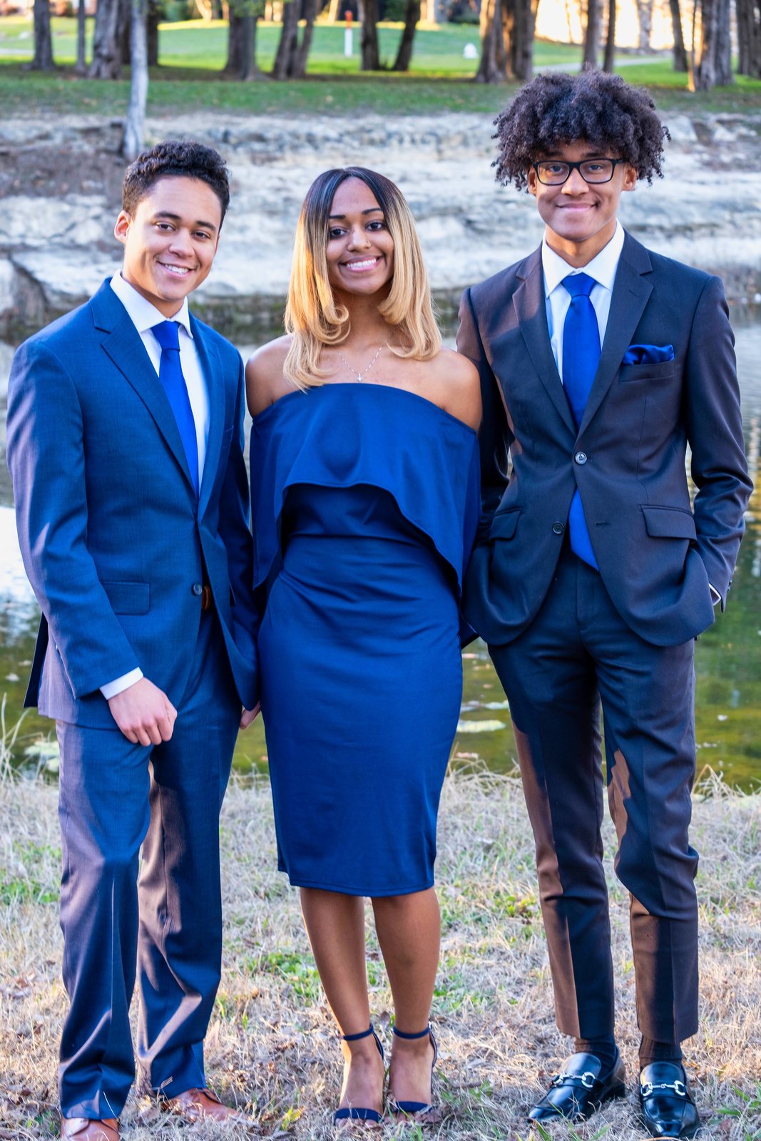 Two young men in navy suits and a woman in a blue dress by a creek
